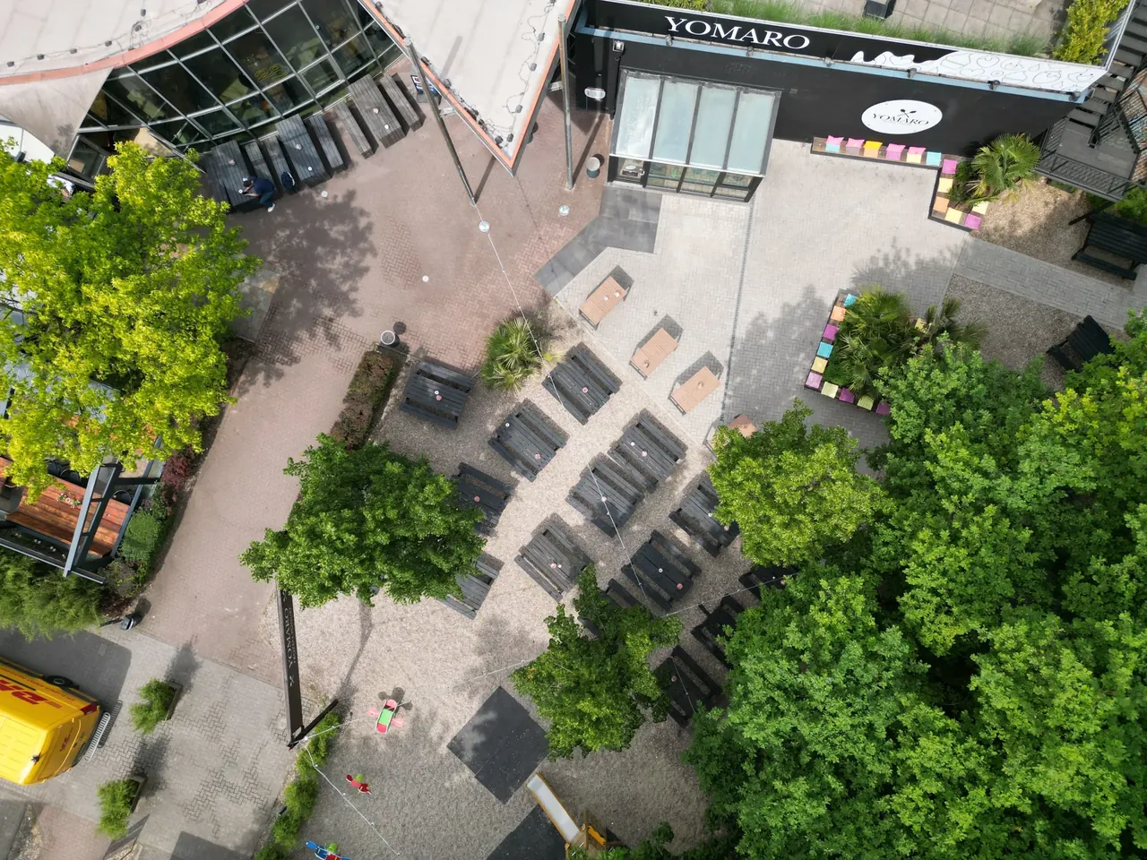 Der Köpi Biergarten senkrecht von oben: Reihen von Bierbänken auf Kies zwischen Baumkronen, oben das YOMARO-Gebäude